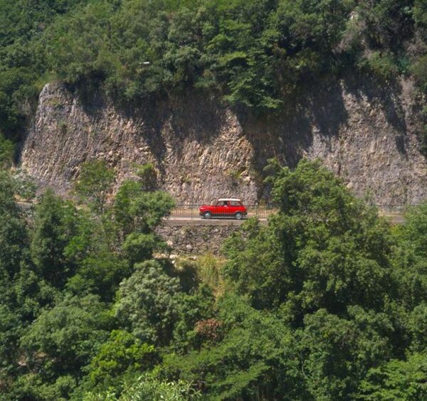 Route d’Agerola avec voiture rouge, falaises rocheuses et végétation luxuriante.
