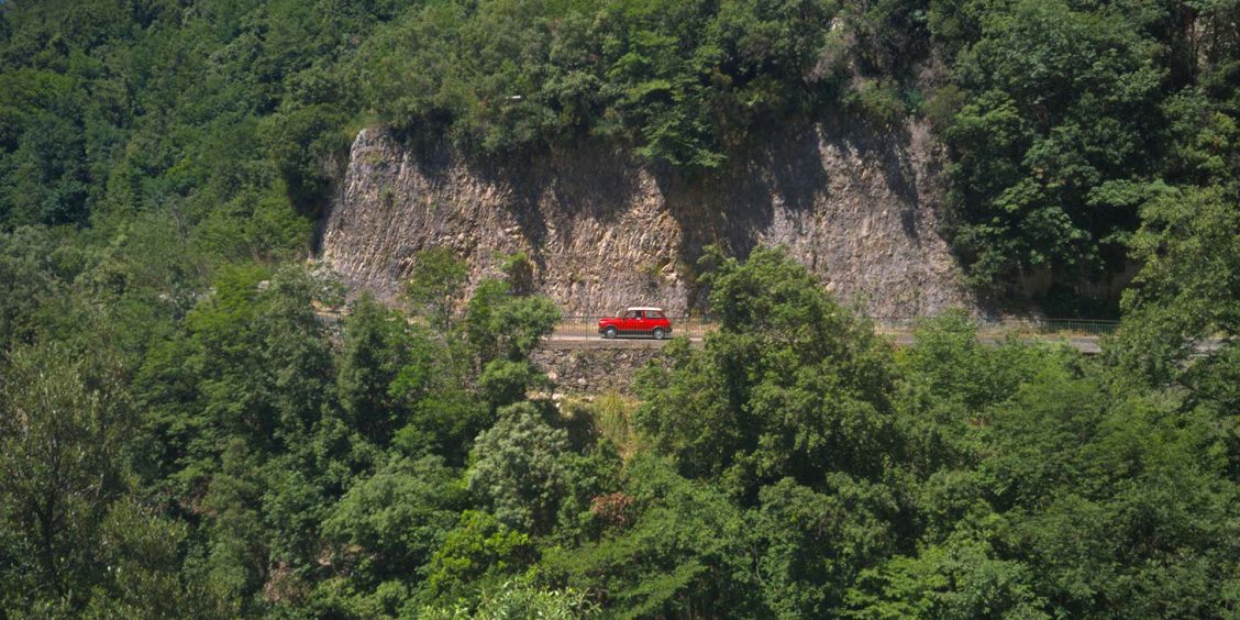 Route d’Agerola avec voiture rouge, falaises rocheuses et végétation luxuriante.