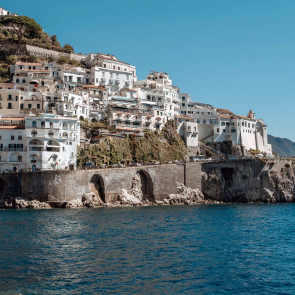 Vue panoramique depuis la mer de la ville d'Amalfi : façades blanches étagées sur la falaise, arcades du front de mer et tour de guet médiévale dominant les eaux azur de la Côte Amalfitaine.