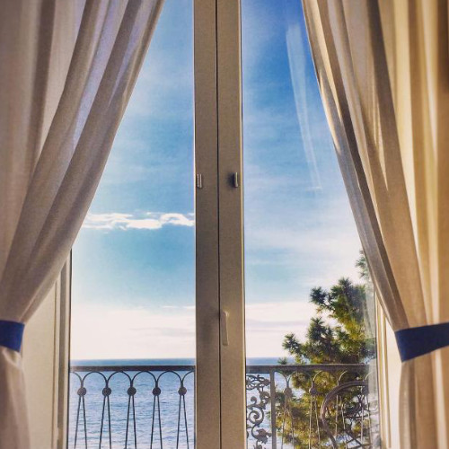 Vue imprenable sur la mer Méditerranée depuis un balcon de chambre encadré par des rideaux ouverts.