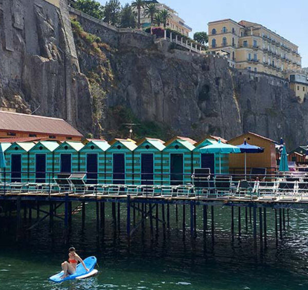 Plage aménagée à Sorrente en août avec cabines colorées, mer et falaises.