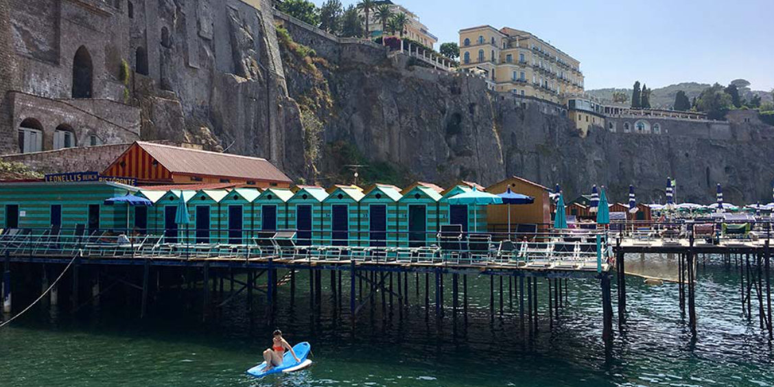 Plage aménagée à Sorrente en août avec cabines colorées, mer et falaises.