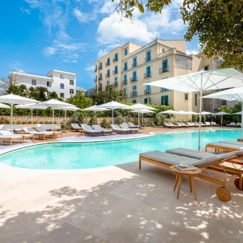 Piscine aux eaux turquoise entourée de nombreux parasols blancs et de transats gris, devant les bâtiments de l'hôtel sous un ciel bleu.