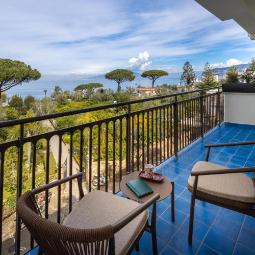 Balcon carrelé de bleu avec un coin salon offrant une vue panoramique sur des jardins arborés et la mer à l'horizon.