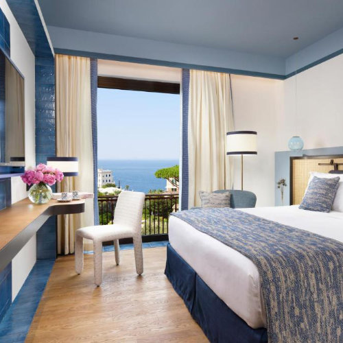 Chambre d'hôtel moderne aux tons bleus et blancs avec un lit confortable et une vue imprenable sur la côte depuis la baie vitrée.
