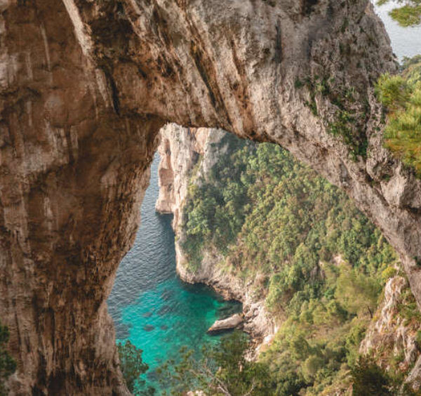 Vue plongeante à travers l'Arco Naturale de Capri sur une mer turquoise et une côte rocheuse sauvage.