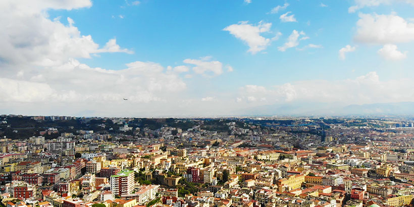 Vue aérienne panoramique de la ville de Naples, ses collines et ses bâtiments sous un ciel bleu parsemé de nuages.