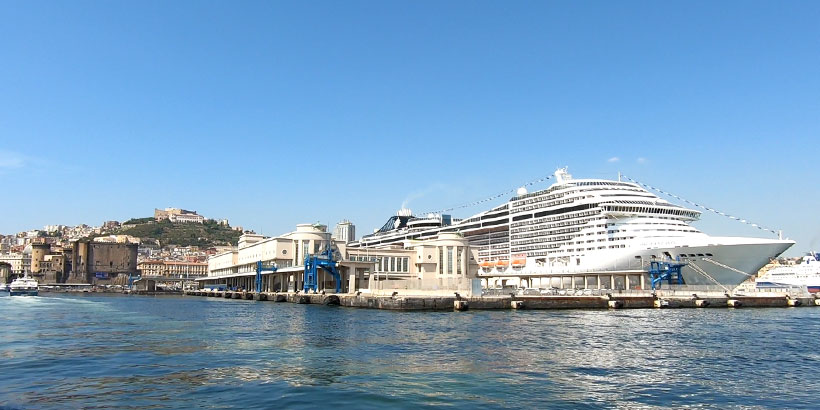 Un imposant paquebot de croisière blanc accosté à la Stazione Marittima de Naples, avec la colline du Vomero en arrière-plan.