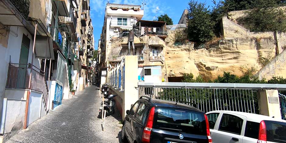 Ruelle napolitaine étroite bordée de voitures en stationnement et d'une imposante paroi de tuf volcanique sous un ciel clair.