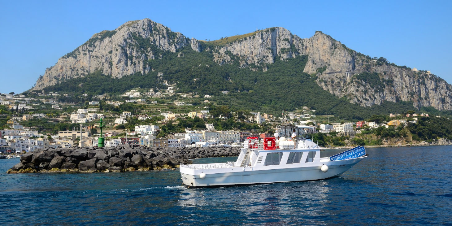 Bateau touristique blanc avec l'inscription « Grotta Azzurra » naviguant devant la côte de Capri dominée par de hautes falaises rocheuses.