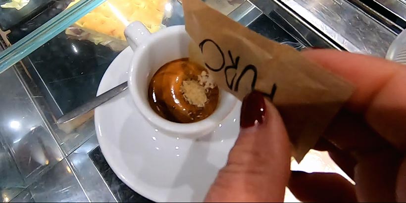 Main tenant un sachet de sucre en train d'être versé dans une petite tasse d'expresso avec une mousse onctueuse, posée sur une soucoupe blanche.