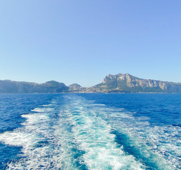 Sillage blanc d'un ferry dans les eaux turquoise de la Méditerranée avec la silhouette de l'île de Capri à l'horizon.