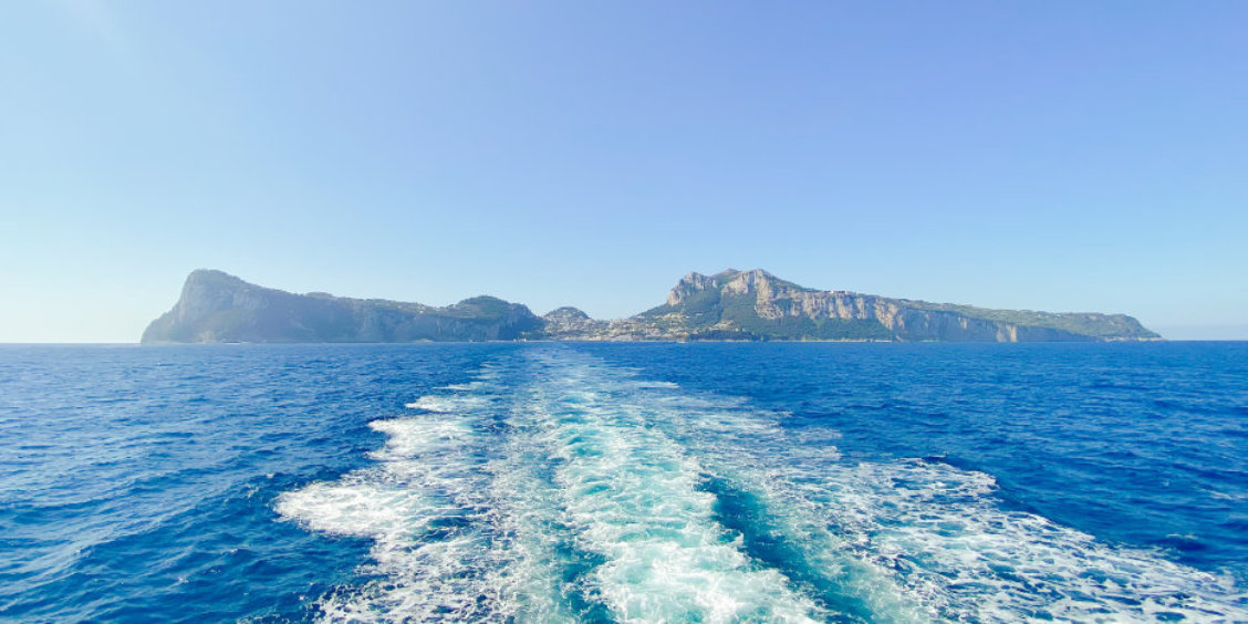 Sillage blanc d'un ferry dans les eaux turquoise de la Méditerranée avec la silhouette de l'île de Capri à l'horizon.