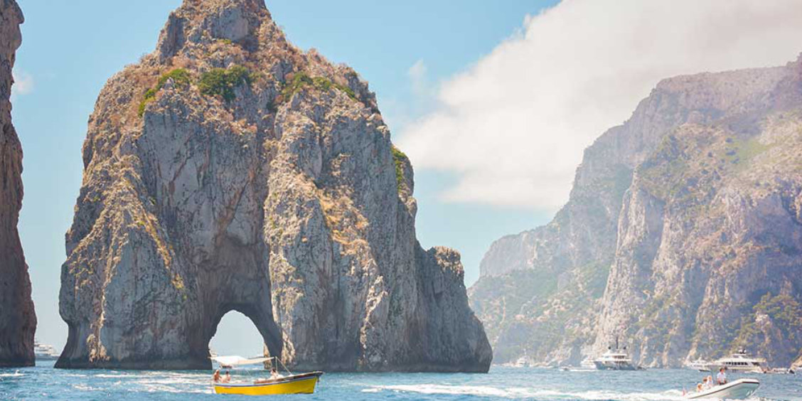 Petit bateau jaune traversant l'arche naturelle des Faraglioni de Capri par une journée ensoleillée.