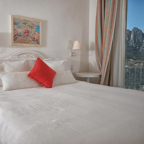 Chambre lumineuse à Capri avec coussin rouge éclatant et vue imprenable sur les montagnes.