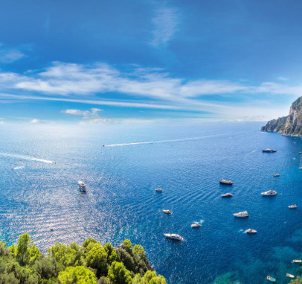 Vue panoramique de la côte de Capri en avril avec les rochers Faraglioni, de nombreux bateaux sur l'eau et des falaises verdoyantes.