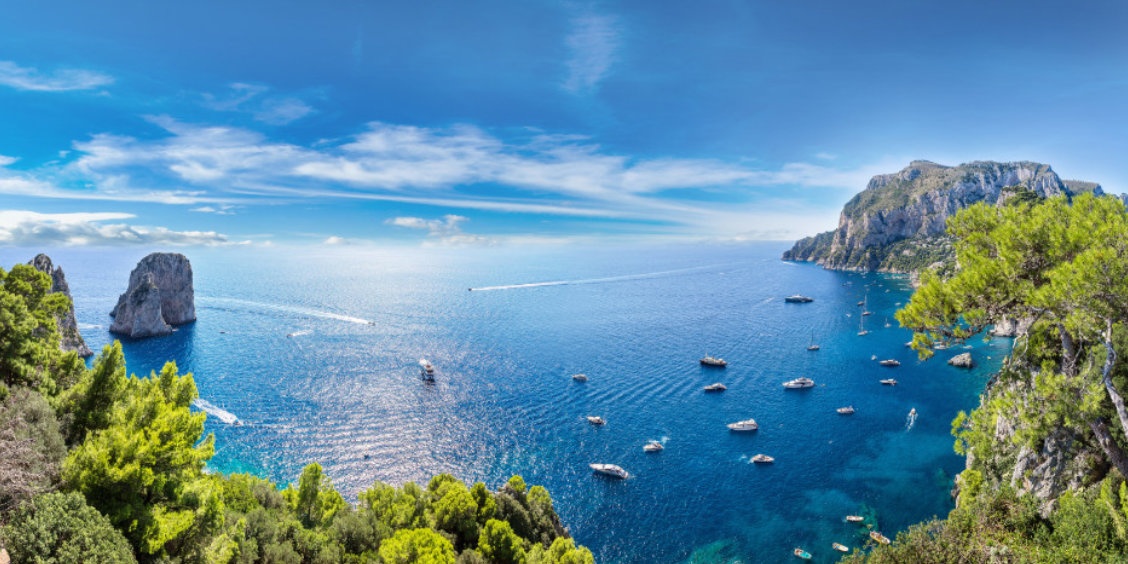 Vue panoramique de la côte de Capri en avril avec les rochers Faraglioni, de nombreux bateaux sur l'eau et des falaises verdoyantes.