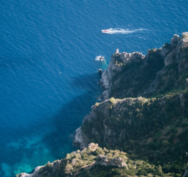 Vue plongeante sur la côte rocheuse de Capri et la mer d'un bleu profond avec un petit bateau au loin.