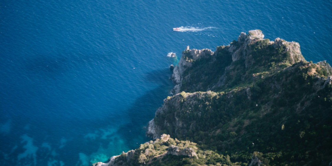 Vue plongeante sur la côte rocheuse de Capri et la mer d'un bleu profond avec un petit bateau au loin.
