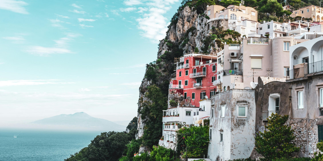 Maisons colorées à flanc de falaise à Capri surplombant la mer avec la silhouette du mont Vésuve à l'horizon.