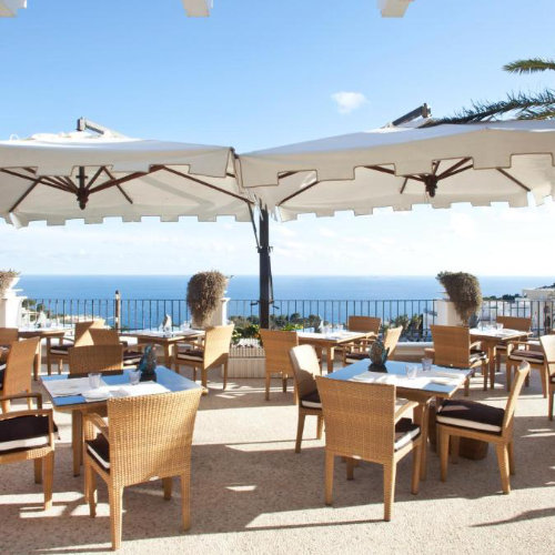 Terrasse du restaurant de l'hôtel Capri Tiberio Palace avec parasols blancs, chaises en osier et vue sur la mer.