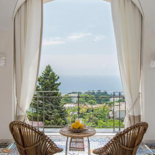 Balcon de l'hôtel Capri Tiberio Palace avec sol en mosaïque, chaises en osier et vue sur la mer à travers une arche.