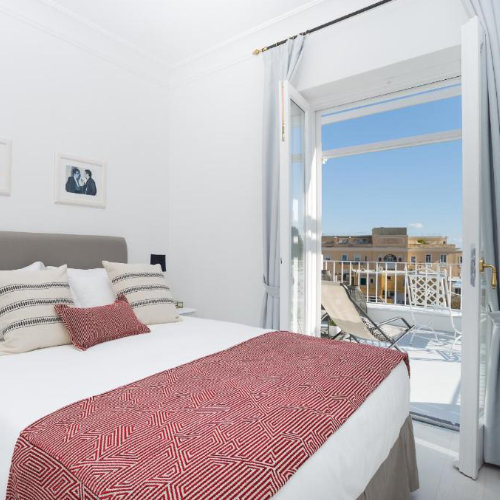 Chambre de la Casa di Capri avec lit blanc, jeté de lit rouge et balcon ouvrant sur une terrasse ensoleillée.