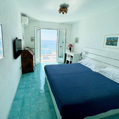 Chambre lumineuse avec sol carrelé bleu et balcon offrant une vue imprenable sur la mer.