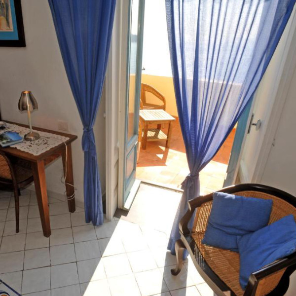 Chambre lumineuse aux rideaux bleus s'ouvrant sulla mer depuis un balcon privé.