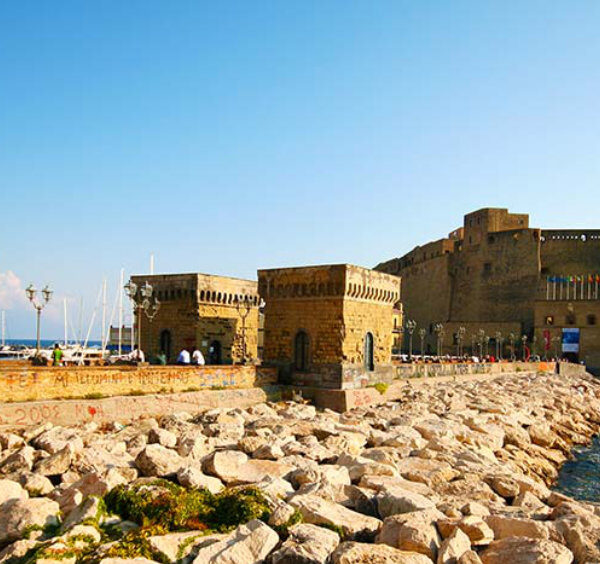Vue du Castel dell'Ovo à Naples depuis le Borgo Marinari avec des bateaux amarrés et un muret en pierre.