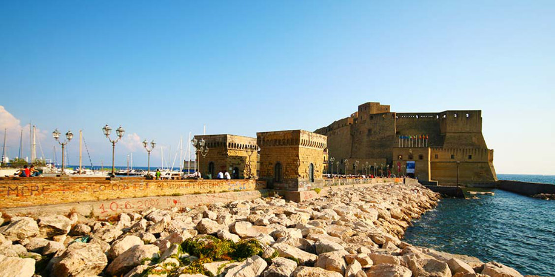 Vue du Castel dell'Ovo à Naples depuis le Borgo Marinari avec des bateaux amarrés et un muret en pierre.