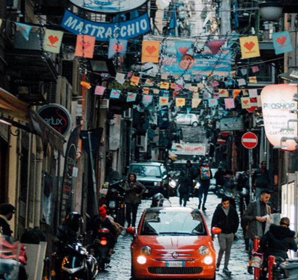 Rue animée du centre historique de Naples avec voitures, piétons et décorations suspendues entre les immeubles.