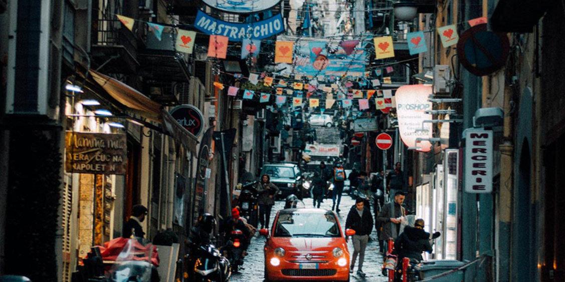 Rue animée du centre historique de Naples avec voitures, piétons et décorations suspendues entre les immeubles.