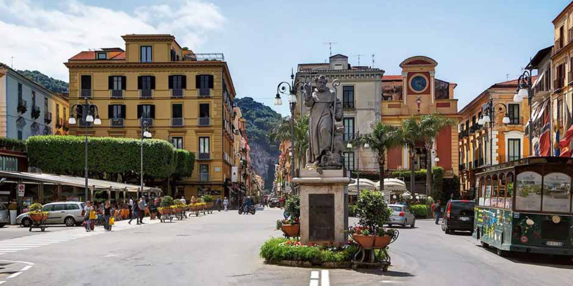 Piazza Tasso à Sorrente avec statue centrale, cafés, bâtiments colorés et petit train touristique.
