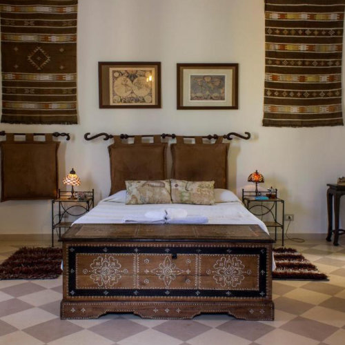 Chambre de charme décorée avec un coffre en bois ancien au pied du lit, des tapisseries ethniques et un sol en damier typique.
