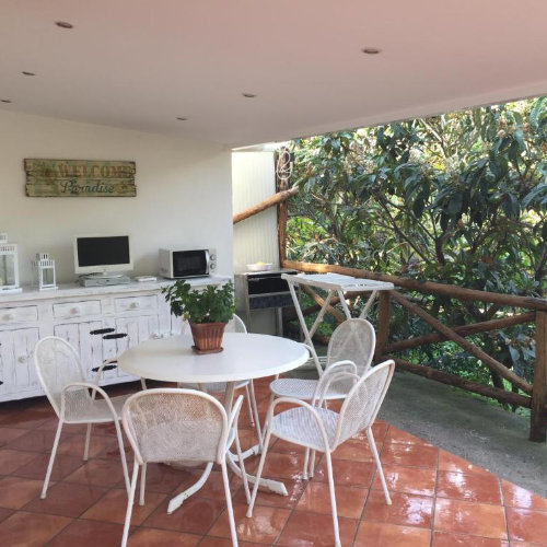 Terrasse couverte d’une location à Sorrente avec table, chaises blanches et jardin ombragé.