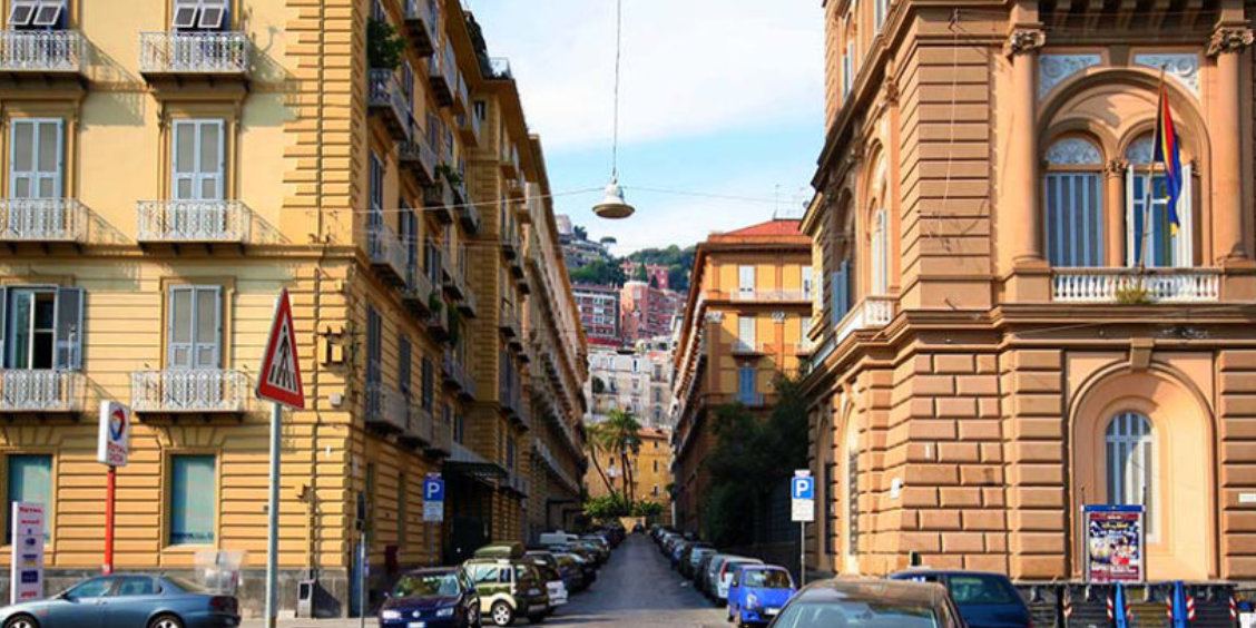 Rue étroite et pavée à Naples bordée d'immeubles historiques colorés, menant vers une colline boisée.