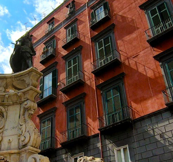 Place animée de Naples avec statue en pierre au premier plan et immeubles colorés aux balcons typiques en arrière-plan.