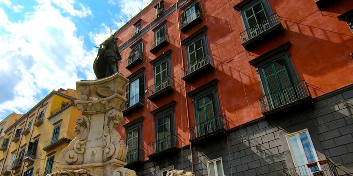 Place animée de Naples avec statue en pierre au premier plan et immeubles colorés aux balcons typiques en arrière-plan.
