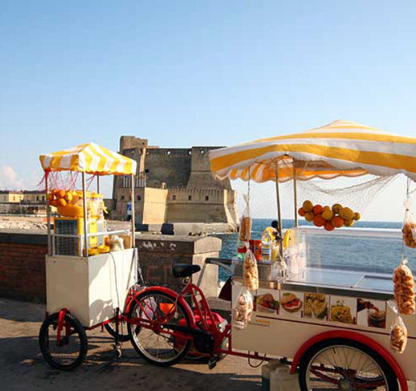 Chariots de vente de produits locaux sur le front de mer de Naples, offrant une vue sur le Castel dell'Ovo et la mer Méditerranée.