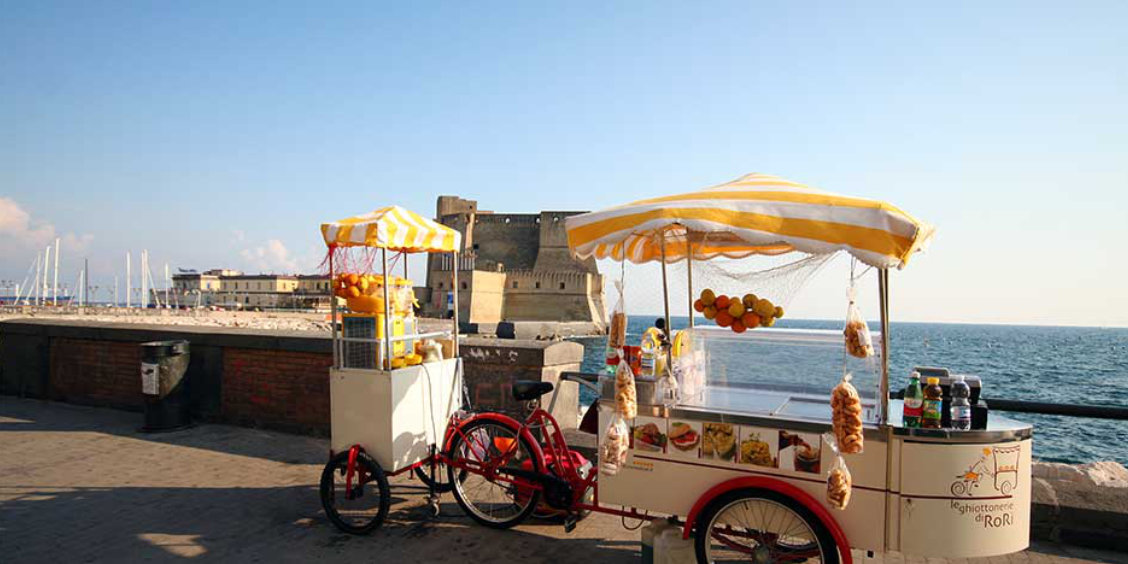 Chariots de vente de produits locaux sur le front de mer de Naples, offrant une vue sur le Castel dell'Ovo et la mer Méditerranée.