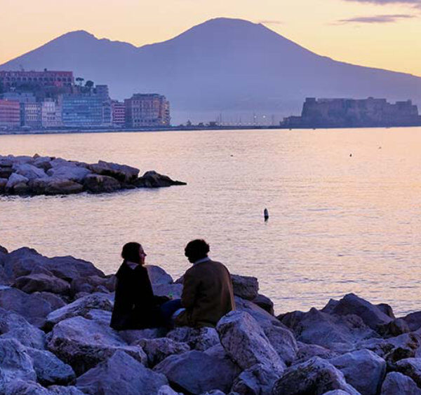 Vue romantique du front de mer de Naples au coucher du soleil avec un couple assis sur les rochers.