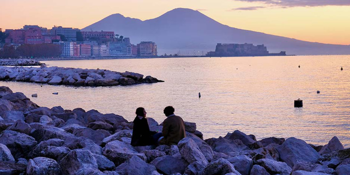 Vue romantique du front de mer de Naples au coucher du soleil avec un couple assis sur les rochers.
