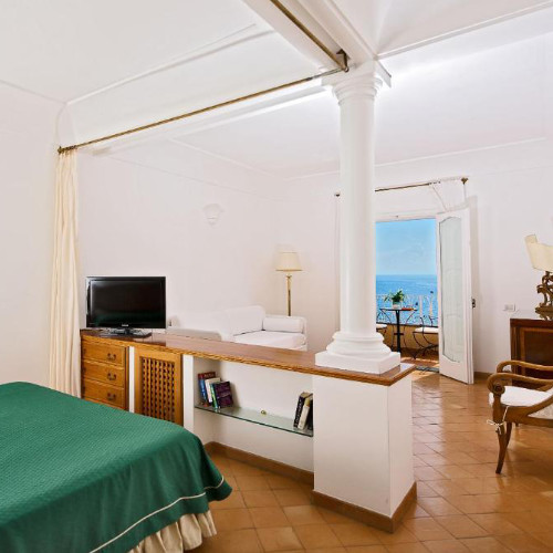 Intérieur d'une suite d'hôtel lumineuse avec carrelage, grand lit, colonne blanche et balcon vue mer.