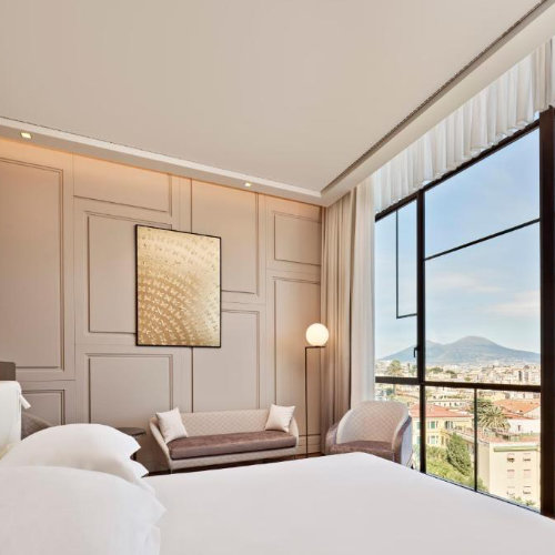 Chambre d'hôtel luxueuse et lumineuse dont la grande fenêtre offre une vue imprenable sur Naples et la silhouette du Vésuve.