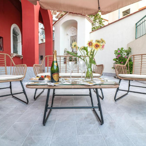 Terrasse accueillante à la Dimora Gioia à Pompéi avec un salon de jardin en rotin, une bouteille de vin pétillant et des fleurs sur la table, devant un mur rouge à arcades.