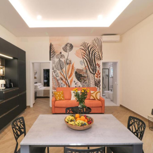 Espace de vie lumineux à la Dimora Gioia avec un canapé orange devant une fresque murale végétale, une cuisine noire et une table de salle à manger au premier plan.