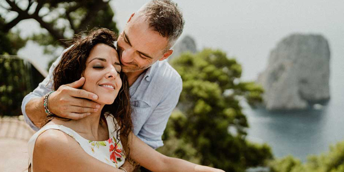 Couple souriant enlacé devant un paysage côtier de Capri avec les rochers Faraglioni en arrière-plan.