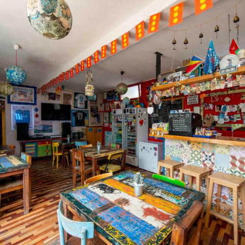 Salle commune colorée et éclectique de l'Eco Hostel Floreale avec des tables en bois recyclé, des murs décorés de souvenirs et un bar.