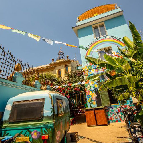 Façade bleu clair de l'Eco Hostel Floreale ornée d'une peinture murale d'arc-en-ciel et de fleurs, avec une caravane peinte dans la cour.